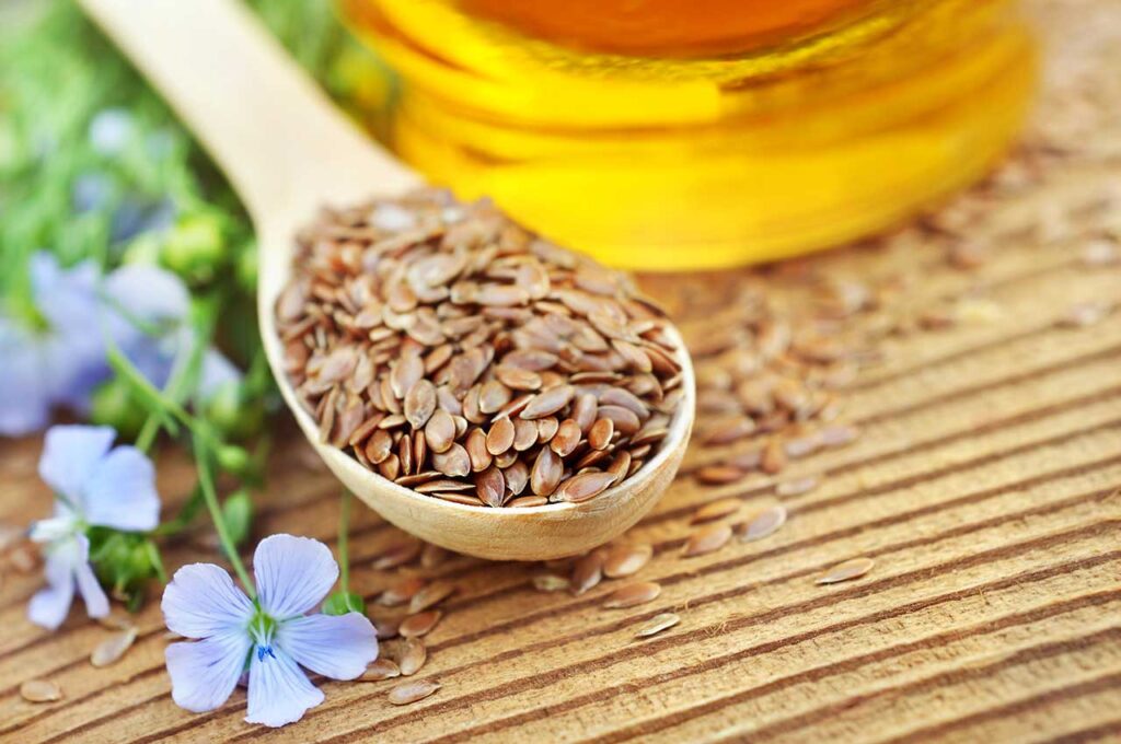 Gesunde Körner ein einem Holzlöffel, im Hintergrund ein Glas mit Öl als Symbol für den Tagesbedarf von Omega-3-Fettsäuren