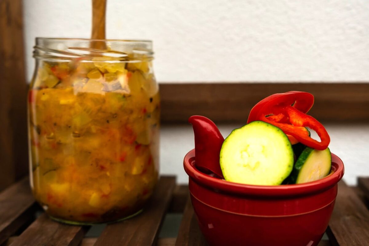 Glas mit Zucchini-Paprika-Relish neben frischen Zutaten in Schale.