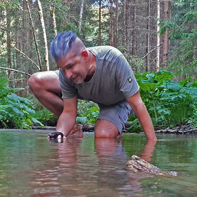 Matthias kniet barfuß in einem klaren Waldbach, beugt sich mit einem Lächeln nach vorne und berührt achtsam das Wasser – umgeben von dichtem Grün, in tiefer Verbindung mit der Natur.