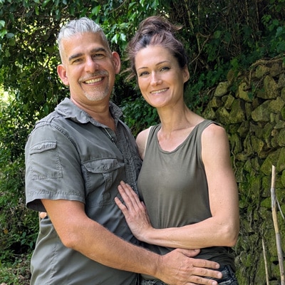 Sandra und Matthias Exl sitzen Arm in Arm am Ufer eines Flusses – ein Moment von Freude, Leichtigkeit und tiefer Zweisamkeit in der Natur.