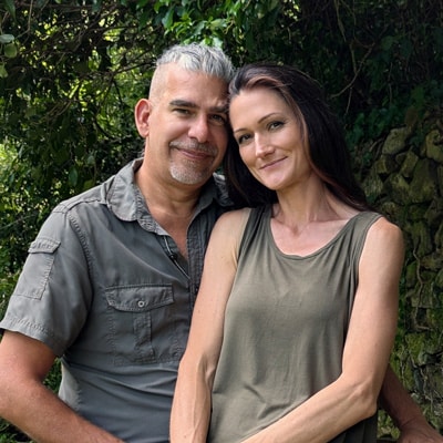 Sandra und Matthias Exl sitzen Arm in Arm am Ufer eines Flusses – ein Moment von Freude, Leichtigkeit und tiefer Zweisamkeit in der Natur.