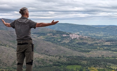 Matthias steht mit ausgebreiteten Armen auf einem Felsvorsprung und blickt in die Weite der portugiesischen Berglandschaft. Im Hintergrund liegt ein kleines Dorf eingebettet zwischen grünen Hügeln und Wäldern. Der Himmel ist bewölkt, die Szene strahlt dennoch Ruhe, Größe und ein tiefes Gefühl von Einklang mit der Natur aus.